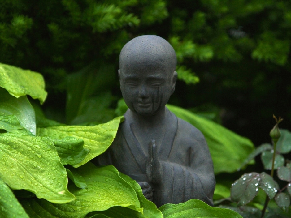 A stone statue of a of a person meditating nestled among lush green leaves.