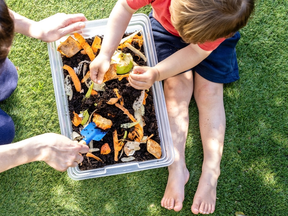 A child's hands breaking up organic food waste into a plastic container filled with dirt and compost material.