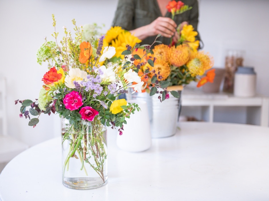 A flower arrangement featuring sunflowers, roses, and other colourful blooms sits in a glass vase on a white table, with a person holding a flower in the background.