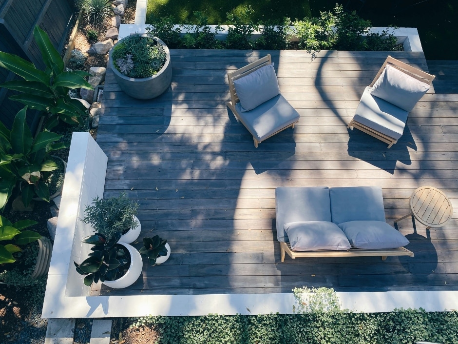 An aerial view of a contemporary outdoor seating area with grey cushioned lounge chairs and potted plants, on a slatted wooden deck.
