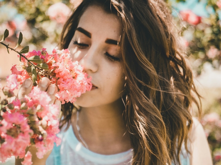 A brown-haired woman smelling the fragrance of a branch of pink flowers.