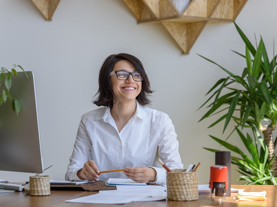 A smiling person wearing glasses and a formal white shirt at an office desk with leaves on the desktop monitor and a green plant in the background.