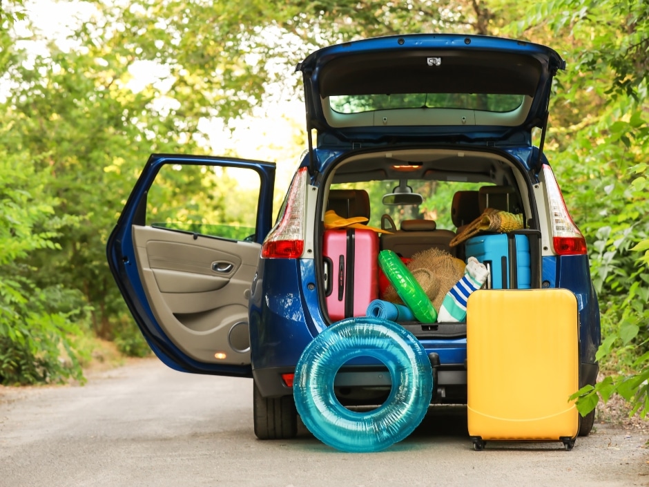 An open car boot packed with luggage, pool toys, and a yellow suitcase, parked on a road amidst greenery.
