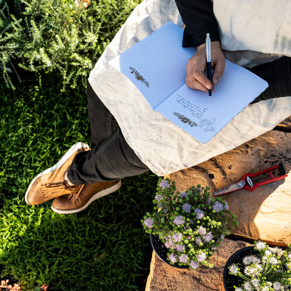 Overhead view of a seated person writing in a notebook, with flowers and gardening tools around them.