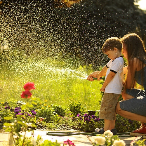 Boy and girl playing with a garden hose spraying water in a sunny, flower-filled yard.