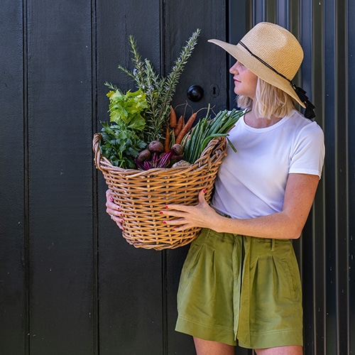 Woman in hat holding wicker basket full of vegetables.