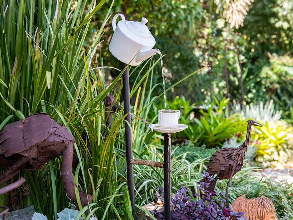 A water feature shaped like a teapot and teacup, surrounded by green plants in a garden setting.