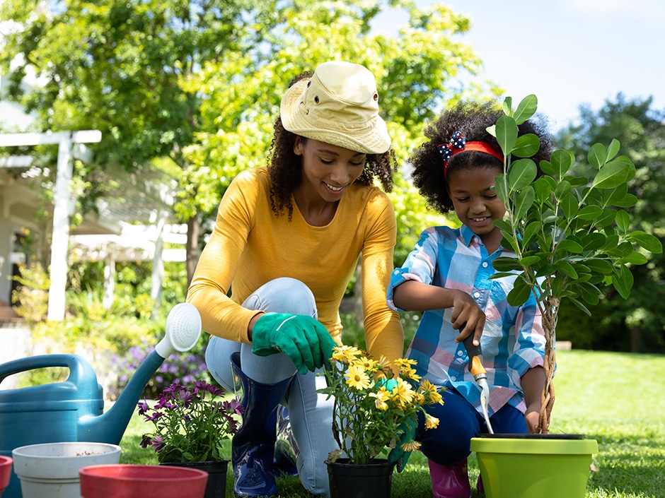 An adult woman and young girl gardening outdoors, wearing hats and gloves while planting flowers in colourful pots on a sunny day.