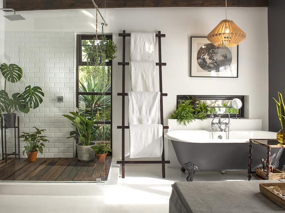 Modern bathroom interior with potted tropical plants, clawfoot bathtub, patterned black and white flooring, and wall decor.