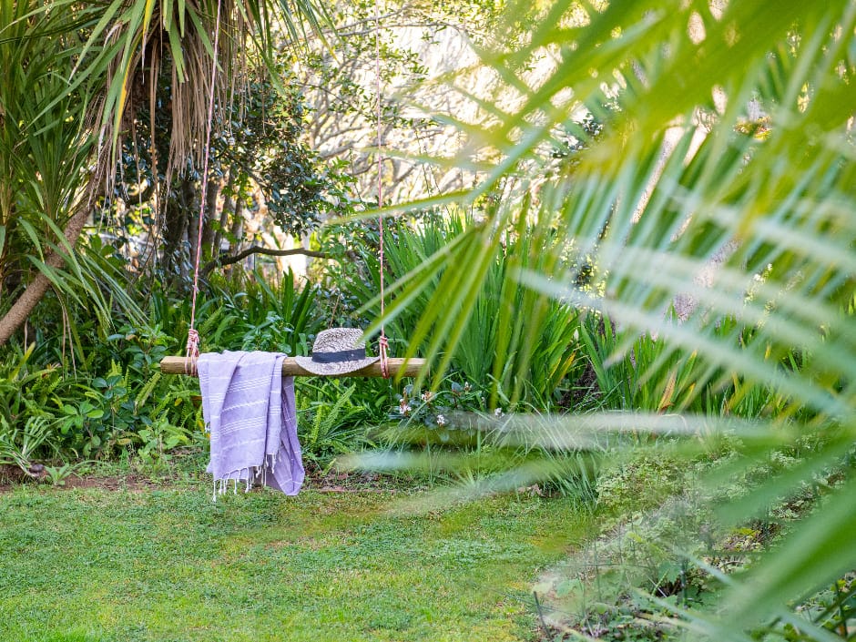 A serene garden with a wooden swing hanging between trees, draped with a towel and a hat, surrounded by lush greenery and soft sunlight filtering through the leaves.