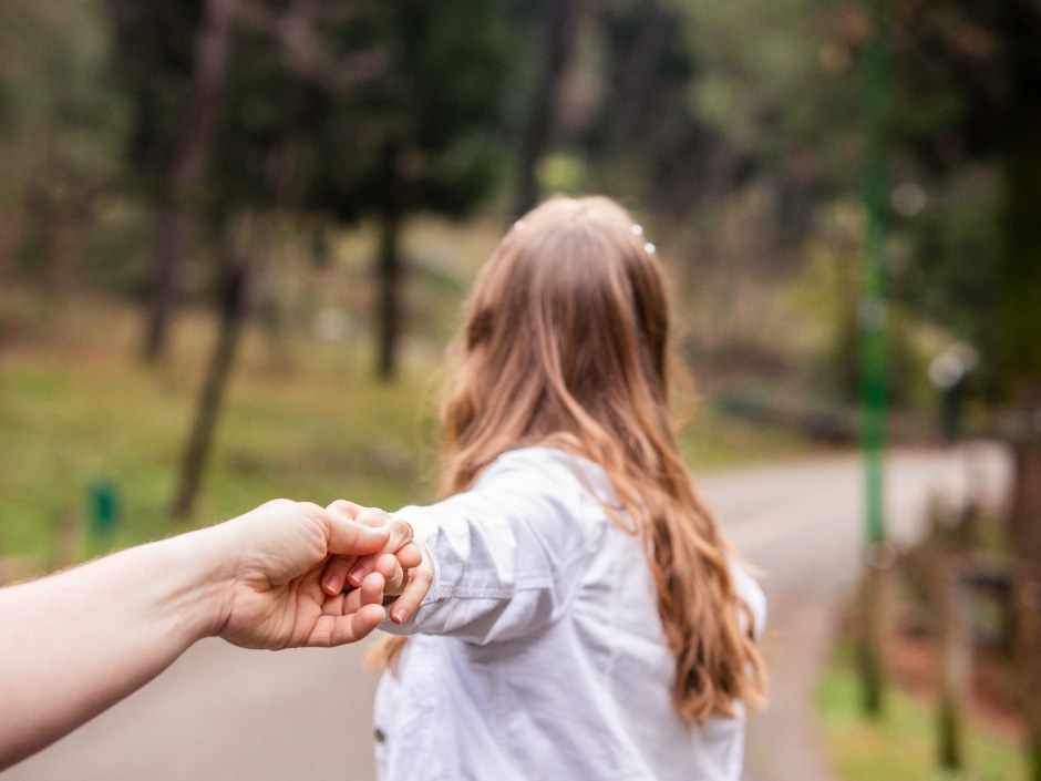 A person's hand holding a woman's hand as she walks away on a path in a park.