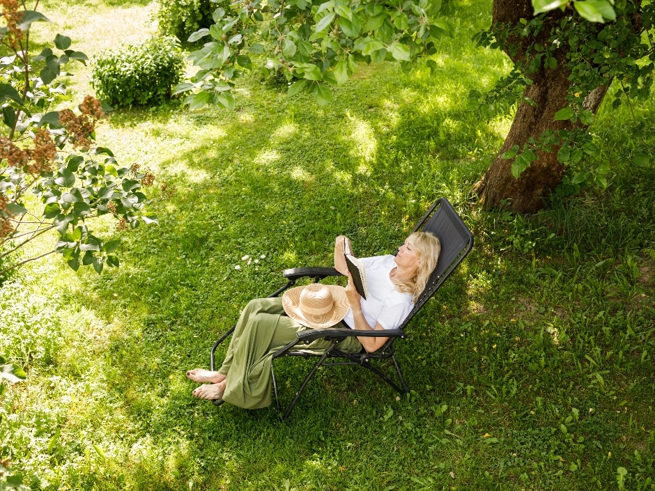 A high-angle view of a woman lounging in a garden chair reading a book in the shade of a large tree, with grass beneath her.