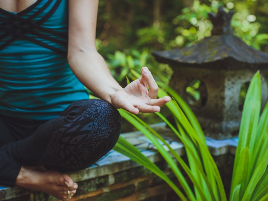 A person sitting in a meditation position in a garden with green plants a moss-covered wall.